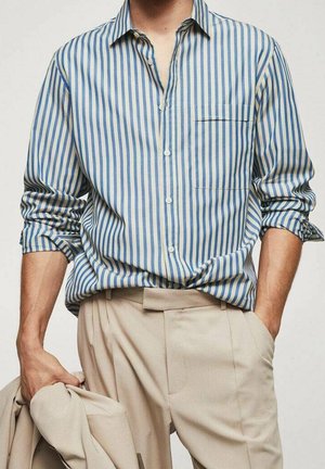 Homme portant une chemise à rayures verticales bleues et blanches avec boutons et manches retroussées, et un pantalon beige, tenant une veste beige dans la main gauche.