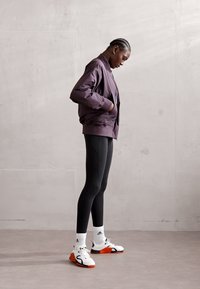 Lila bomber dzseki, fekete leggings és fehér sportcipő narancs részletekkel. A alany oldalról áll egy semleges háttér előtt.