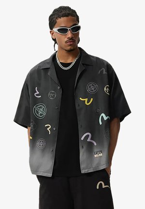Uomo con camicia nera con bottoni con simboli astratti colorati, t-shirt nera e pantaloncini, collana con catena d'argento e occhiali da sole bianchi.