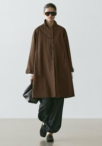 Woman walking wearing oversized brown buttoned coat, black loose pants, black loafers, dark sunglasses, and carrying a black handbag.