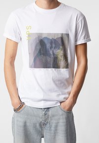 Bílý tričko s předním potiskem, který zobrazuje abstraktní postavy v jemných barvách, doplněné žlutým písmem a horizontálním textem na spodní části.