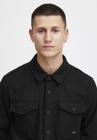 Black button-up jacket features a collar, two chest pockets with flap closures, and a soft texture. Buttons are round and black.