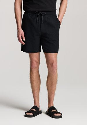 Mann trägt schwarze Shorts mit Kordelzug und schwarze offene Sandalen, steht mit den Händen in den Taschen vor einem einfarbigen Hintergrund.