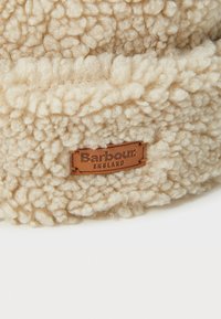 Béžová nadýchaná čepice vyrobená z texturovaného fleecu s koženým štítkem označeným "Barbour England" připojeným na přední straně.