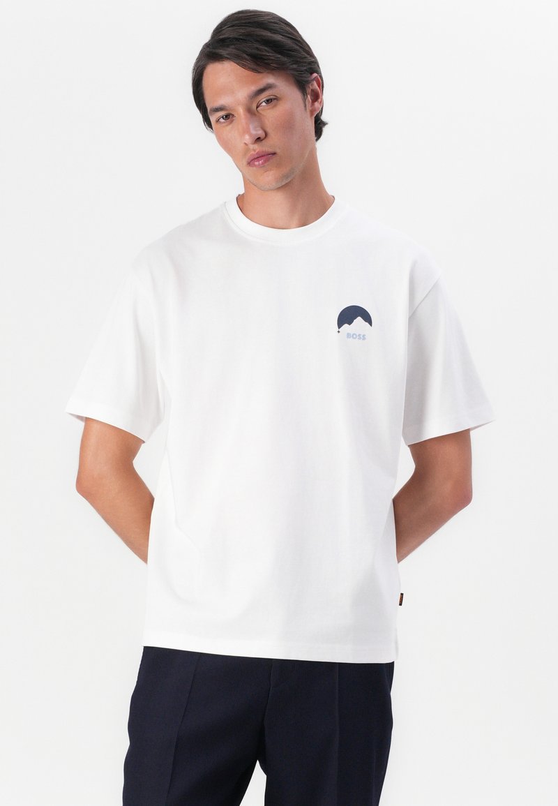T-shirt en coton blanc à manches courtes, avec un motif de logo bleu marine sur le côté gauche de la poitrine. Encolure ronde, coupe décontractée, style minimaliste.