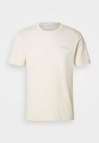 Camiseta de algodón de color crema, con mangas cortas y cuello redondo, con un pequeño logo de "Calvin Klein" en marrón claro en el pecho. Textura suave.