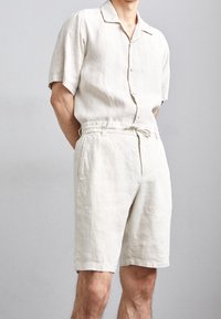 Camicia a maniche corte in lino beige chiaro con colletto e bottoni, abbinata a pantaloni corti in lino beige con vita elastica e tasche laterali.