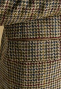 Detailný záber na látku vlneného kabáta s multicolor houndstooth a károvaným vzorom, ktorý obsahuje horizontálnu kapsu s koženým lemovaním.