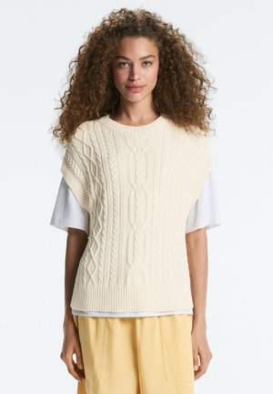 Jonge vrouw met krullend haar met een crème kabelgebreide mouwloze trui over een wit T-shirt en gele broek, staand tegen een witte achtergrond.
