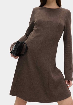 Femme portant une robe marron à manches longues tenant un petit sac à main noir devant un fond uni.