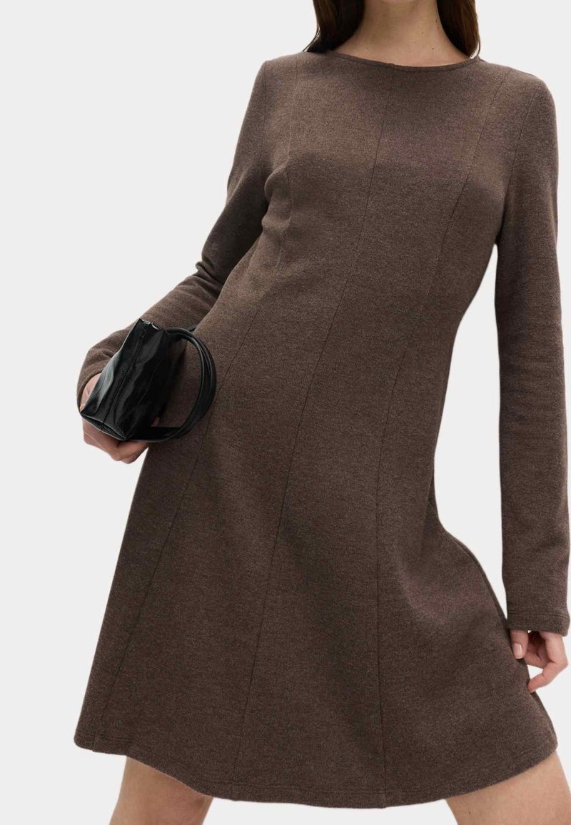 Femme portant une robe marron à manches longues tenant un petit sac à main noir devant un fond uni.