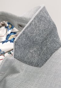 Gros plan d'un col de manteau gris clair avec un sous-col en feutre gris plus foncé et une doublure à motif floral dans des tons bleus et crème.