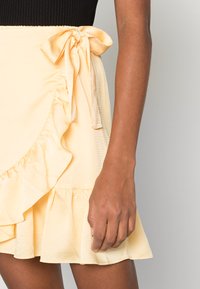 Saia envolvente amarela com bainha franzida e detalhe de laço. Feita de um tecido texturizado com uma sensação leve e arejada e um brilho subtil.