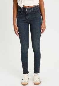 Schmale Jeans aus dunkelblauem Denim, hoch geschnitten mit zwei Knöpfen, fünf Taschen und dezenten Stickdetails. Kombiniert mit weißen Sneakers.