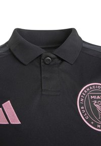 Juoda polo marškinėliai su sagotu apykakle, kairėje krūtinės pusėje rožinis Adidas logotipas, dešinėje krūtinės pusėje rožinis Majamio futbolo klubo emblem.