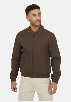 Mężczyzna ubrany w brązową kurtkę na zamek z kołnierzem i elastycznymi mankietami, w połączeniu z jasnobeżowymi spodniami, patrzący lekko w prawo.