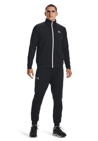 Zwarte sportieve trainingspak gemaakt van gladde stof, met een zipjack met hoge kraag en bijpassende slimfit broek. Under Armour-logo zichtbaar.