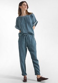 Donna che indossa una maglia blu con maniche arrotolate e pantaloni abbinati con coulisse, scarpe basse bordeaux con lacci, in piedi davanti a uno sfondo bianco.