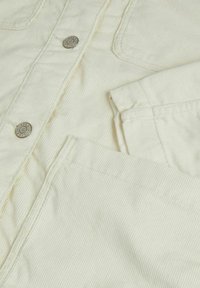 Close-up of off-white corduroy jacket showing metal buttons, a folded sleeve, and stitched pockets.