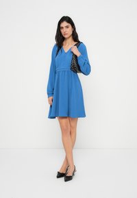 Μπλε φόρεμα με μακριά μανίκια, V-λαιμόκοψη, τονισμένη μέση και φαρδύ φούστα. Συνδυασμένο με μαύρη τσάντα με πουά και μαύρα μυτερά παπούτσια.