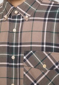 Ruskea ruudullinen paita, jossa vihreitä, mustia ja valkoisia kuvioita, nappikiinnitteinen kaulus ja rintatasku, valmistettu teksturoidusta kankaasta.