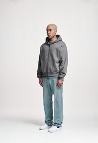 Szara bluza z kapturem zapinana na zamek, z dużym kapturem, dwoma przednimi kieszeniami, noszona na jasnej koszulce, w zestawieniu z jasnoniebieskimi dżinsami i białymi trampkami.