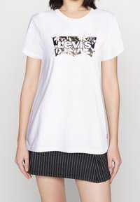 Bílá bavlněná tričko s černým a květinovým logem Levi's. Tričko má kulatý výstřih a krátké rukávy, vystavené s pruhovanou sukní.