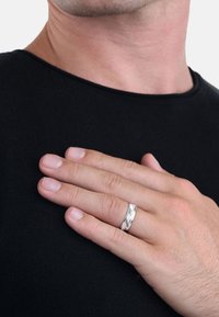 Anello in metallo argento con un motivo intrecciato, esposto su una mano appoggiata su una camicia nera. Texture liscia, finitura lucida e design moderno.
