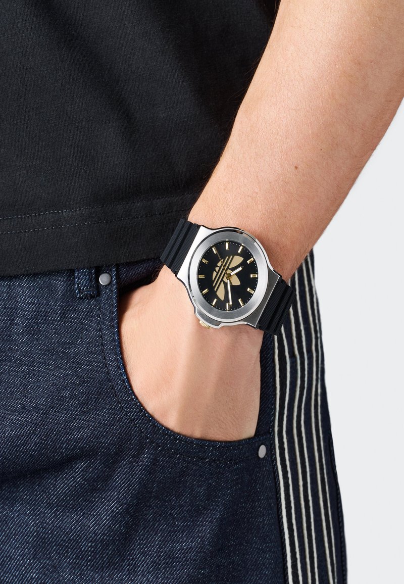 Riešinis laikrodis su juodu ciferblatu ir auksiniais žymekliais ant riešo, ranka tamsiai mėlynos džinsinės kišenės viduje, žmogus vilkintis juodą marškinį.