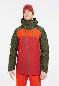 Rode en groene geïsoleerde winterjas met een capuchon, voorzien van ritszakken en een bicolor ontwerp. Draagtextiel is een oranje beanie.
