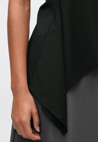 Lewa ręka z naturalnymi paznokciami spoczywa obok czarnej asymetrycznej bluzki i szarej spódnicy lub spodni.