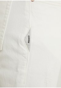 Tissu en denim blanc avec une petite étiquette triangulaire de poche portant l'inscription "CHASIN'" en texte noir près de la couture cousue et d'un rivet en cuivre.