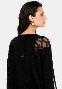 Felpa nera con dettagli in pizzo sulle spalle, scollatura rotonda e logo dorato sul retro. Presenta una vestibilità comoda e una texture morbida.