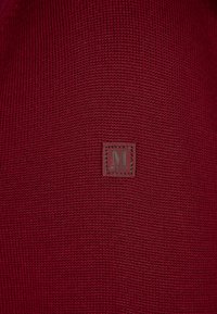 Nahaufnahme eines maroonfarbenen, gestrickten Stoffes mit einem kleinen quadratischen Lederpatch, das in der Mitte mit dem Buchstaben "M" und dem Text "MRZ" bestickt ist.