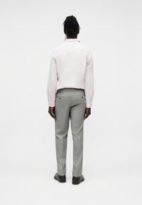 Personne à la peau sombre et aux cheveux tressés, vue de dos, portant une chemise blanche à manches longues, un pantalon gris à fines rayures et des chaussures noires sur fond blanc.