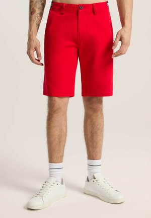 Czerwone bawełniane szorty o prostym kroju, z przednimi kieszeniami i zapięciem na guzik. Noszone z białymi trampkami i paskowanymi skarpetkami.