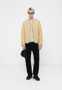 Lys beige dunjakke med glidelås, grå hettegenser under, svarte bukser og sneakers. Blåtonede solbriller og en svart caps holdt i hånden.