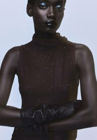 Top negru fără mâneci cu un gât înalt, realizat dintr-un material texturat presărat cu mici accente strălucitoare. Modelul poartă mănuși negre lungi.