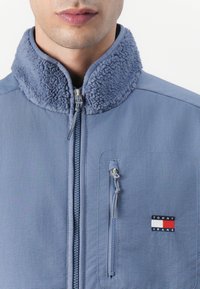 Giacca azzurro chiaro con colletto peloso, zip frontale e una piccola tasca. Presenta il logo "Tommy Jeans" e un design del tessuto testurizzato.