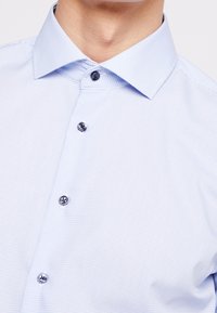 Vaaleansininen nappikauluspaita ruutukuvioinnilla, siistillä kauluksella ja tummilla napeilla. Valmistettu sileästä kankaasta, jossa on hienovaraista tekstuuria.