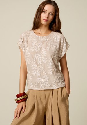 Kvinde iført beige blomstrede blonde kortærmet top, løse brune bukser og chunky røde og gyldne armbånd på venstre håndled, stående med hånd i lommen.