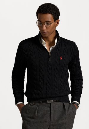 Black cable-knit pullover with a half-zip collar, featuring a red embroidered logo on the chest and ribbed cuffs and hem. Paired with gray trousers.