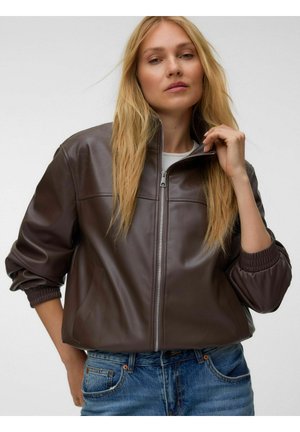 Frau mit langen blonden Haaren, die eine braune Lederjacke mit Reißverschluss und blaue Jeans trägt, hält den Jackenkragen mit einer Hand.