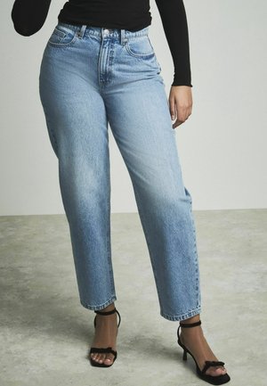 Kvinde iført lyseblå højtaljede jeans, sort langærmet top og sorte hælede sandaler på en grå gulv.