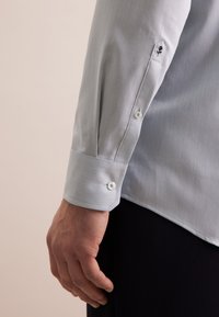 Camicia blu chiaro con finitura testurizzata, dotata di polsini con bottone e un piccolo logo vicino alla manica. Il materiale sembra liscio.