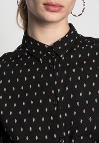 Černá košile na knoflíky s malým krémovým vzorem listů. Tkanina vypadá měkce, má hladkou texturu a klasický design límce.