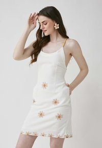 Weißes Kleid mit Sonnenmotiven in verbranntem Orange, dünnen gelben Trägern, tailliertem Schnitt und Seitentaschen. Glattes Material und knielanger Schnitt.