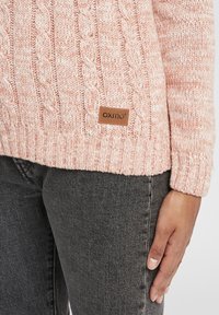 Pink knitted sweater with a cable design, textured finish, and a brown logo tag at the hem. Worn over gray jeans.