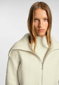 Vit texturerad jacka med hög krage och dragkedjestängning, lager på lager över en ljuskrämfärgad ribbad tröja. Slät vadderad tyg med mjukt avslut.