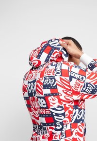 Hombre ajustando la capucha de una sudadera roja, blanca y azul cubierta con los logotipos repetidos de Coca-Cola y Tommy Jeans sobre un fondo blanco liso.
